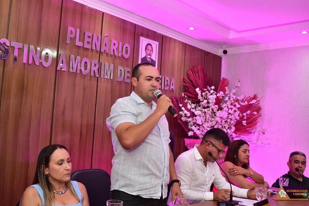Homem em pé fala ao microfone em um plenário com painel de madeira ao fundo, onde se lê “PLENÁRIO AGOSTINHO AMORIM DE ...”. Ele veste camisa clara e está ao lado de uma mesa com outras quatro pessoas sentadas, que observam ou escrevem. Ao fundo, há um arranjo decorativo com flores brancas e folhas vermelhas, sob iluminação rosada.