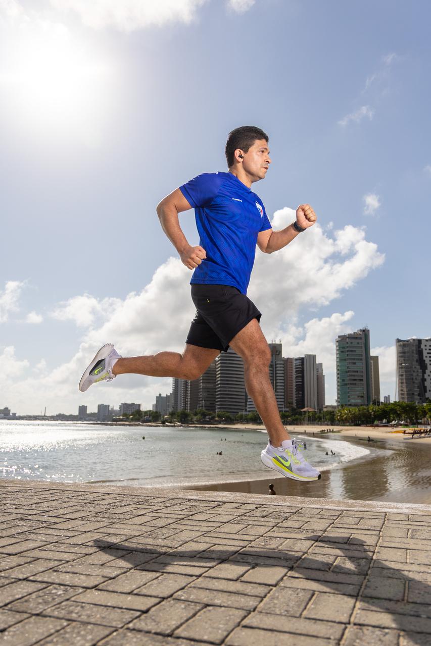 Homem de camiseta azul e bermuda preta correndo em uma área de beira mar.