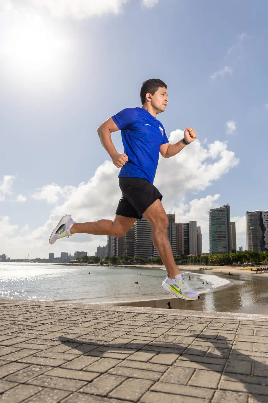 Homem de camiseta azul e bermuda preta correndo em uma área de beira mar.