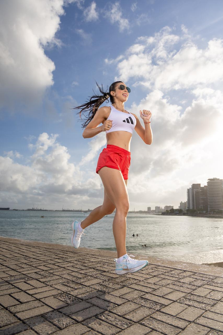 Jovem de cabelo comprido preto, preso em rabo de cavalo, com top branco e short vermelho, correndo em um espigão da Beira Mar de Fortaleza.
