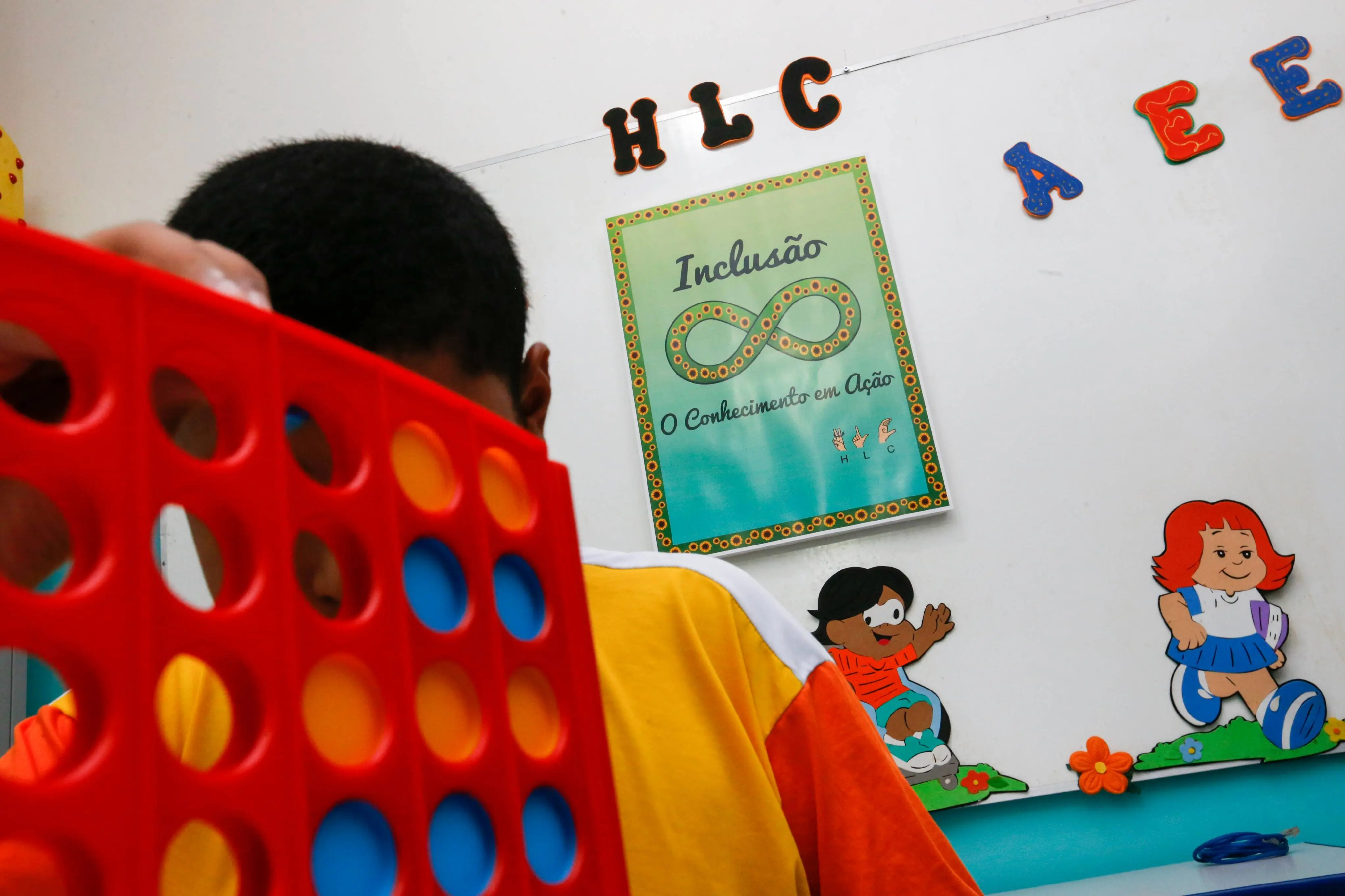 Criança em sala de Atendimento Educacional Especializado segura um brinquedo de encaixe com peças coloridas. Ao fundo, cartaz com o símbolo do infinito e a frase “Inclusão: o conhecimento em ação”, além de ilustrações de crianças e letras decorativas na parede.