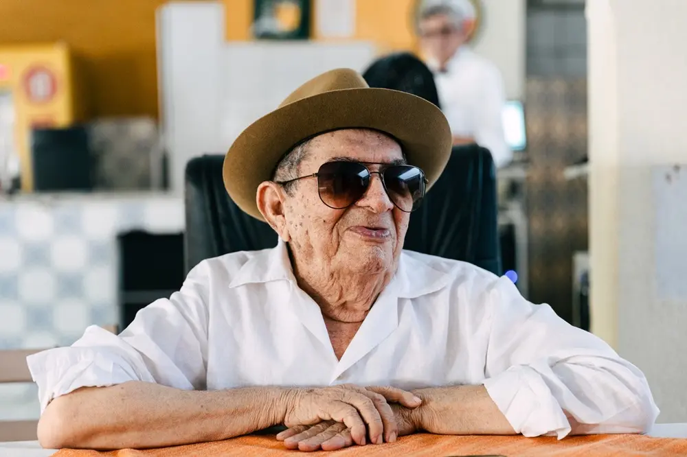 Na imagem, um senhor idoso, de pele clara, está sentado à mesa, olhando para o lado direito. Ele usa uma camisa branca de linho, um chapéu Fedora marrom e óculos escuros de sol. Suas mãos estão cruzadas e repousadas sobre uma toalha de mesa laranja ou um descanso de mesa. No fundo, desfocado, vê-se outra pessoa de camisa branca. A imagem tem a luz natural de um ambiente interno bem iluminado ou externo coberto.