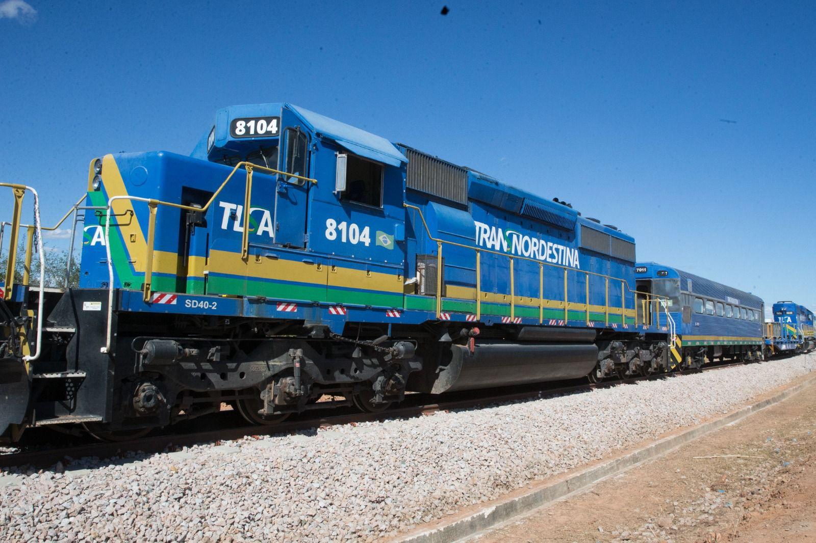 Trem na cor azul, com detalhes em amarelo e verde, em uma linha de ferrovia, com céu azul ao fundo e cascalho no chão.