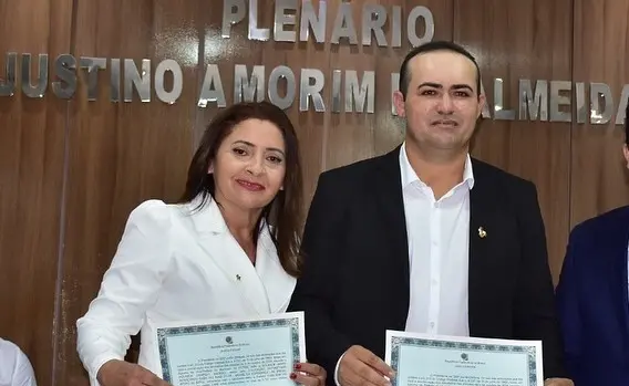 Duas pessoas posam para foto em um plenário com parede de madeira e letreiro metálico ao fundo que diz “PLENÁRIO JUSTINO AMORIM DE ALMEIDA”. Ambas seguram certificados: uma mulher vestida de branco, à esquerda, e um homem de terno preto e camisa branca, à direita. Eles sorriem levemente para a câmera. São Solange Campelo e Luan Dantas.