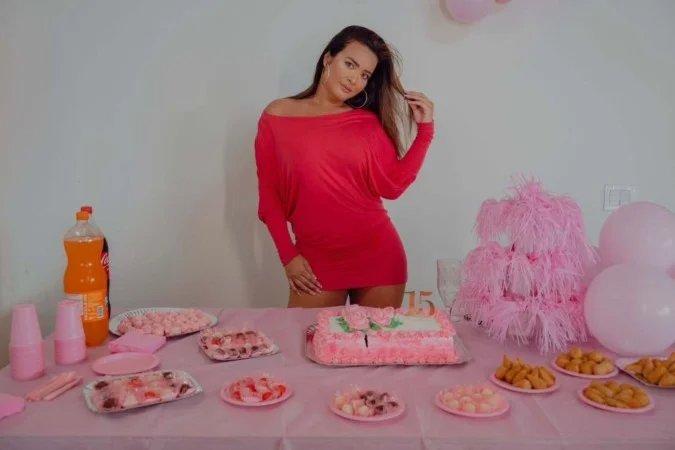 A imagem mostra uma Geisy Arruda posando em frente a uma mesa decorada para uma festa. Ela está usando um vestido curto rosa.