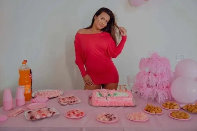 A imagem mostra uma Geisy Arruda posando em frente a uma mesa decorada para uma festa. Ela está usando um vestido curto rosa.