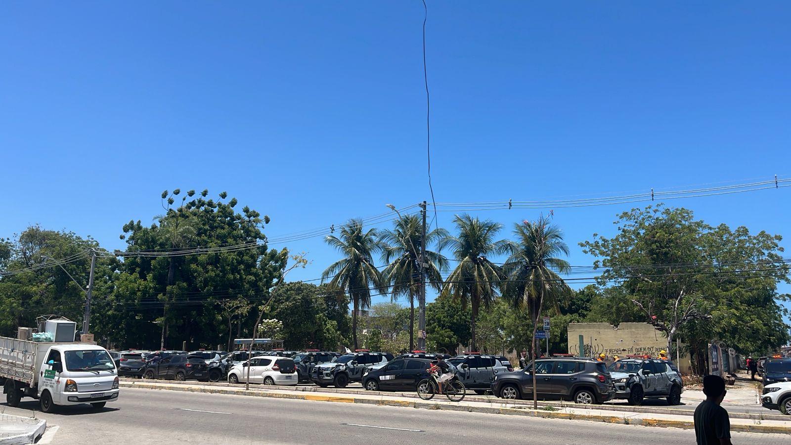 Imagem mostra mais de 10 viaturas policiais estacionadas em frente a um terreno de escola estadual desocupado, cheio de árvores e com céu azul, no bairro Monte Castelo, em Fortaleza. Na avenida em frente, há um fluxo intenso de veículos. A abordagem buscou desocupar uma invasão no local.