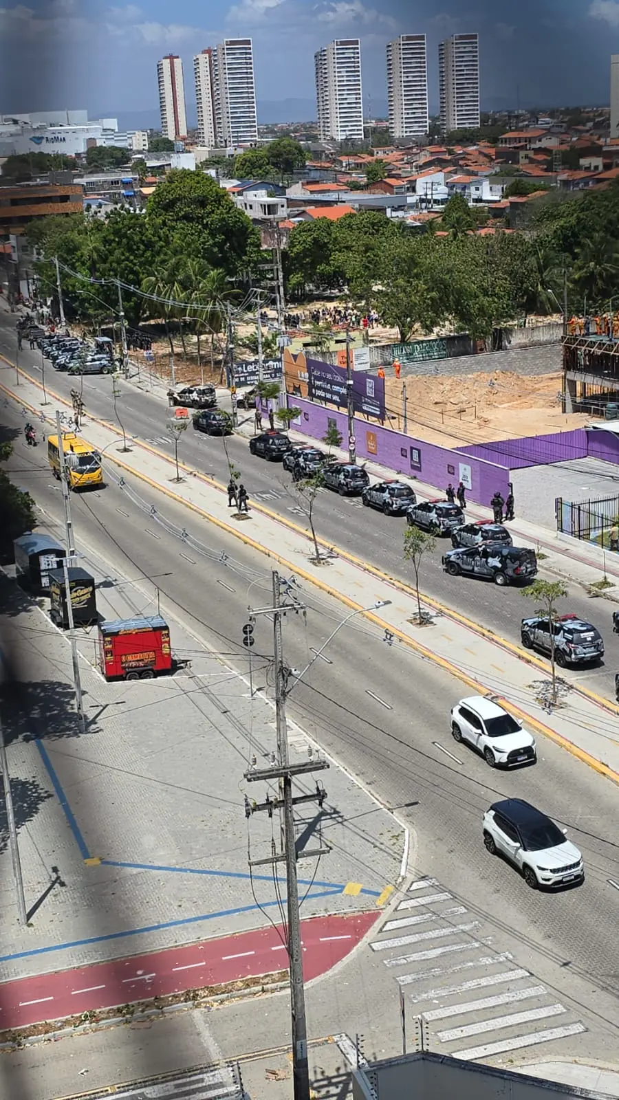 A imagem mostra uma vista aérea da Avenida Sargento Hermínio, com várias viaturas policiais alinhadas ao longo da pista e agentes em operação. À esquerda, um ônibus amarelo circula e, no canto inferior, há um carrinho vermelho de lanches parado na calçada. Do outro lado da via, vê-se um terreno cercado por um muro roxo e uma aglomeração de pessoas próxima a uma área arborizada. Ao fundo, destacam-se prédios altos e o contraste entre construções verticais e casas de telhados baixos.
