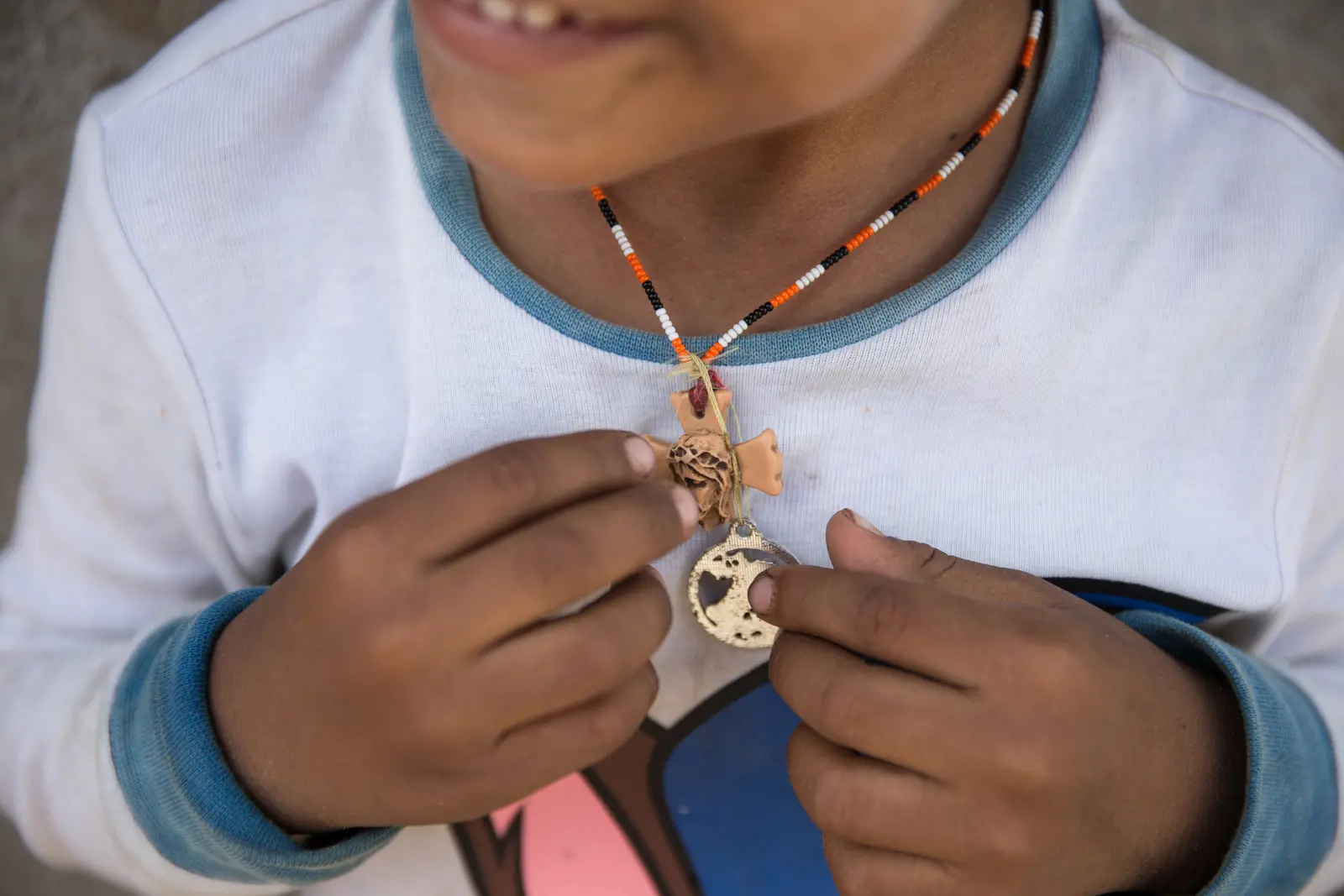 Imagem mostra uma criança da etnia Warao em Maracanaú, no Ceará. Aparecem parte do sorriso do menino e as mãos mostrando um colar de contas.