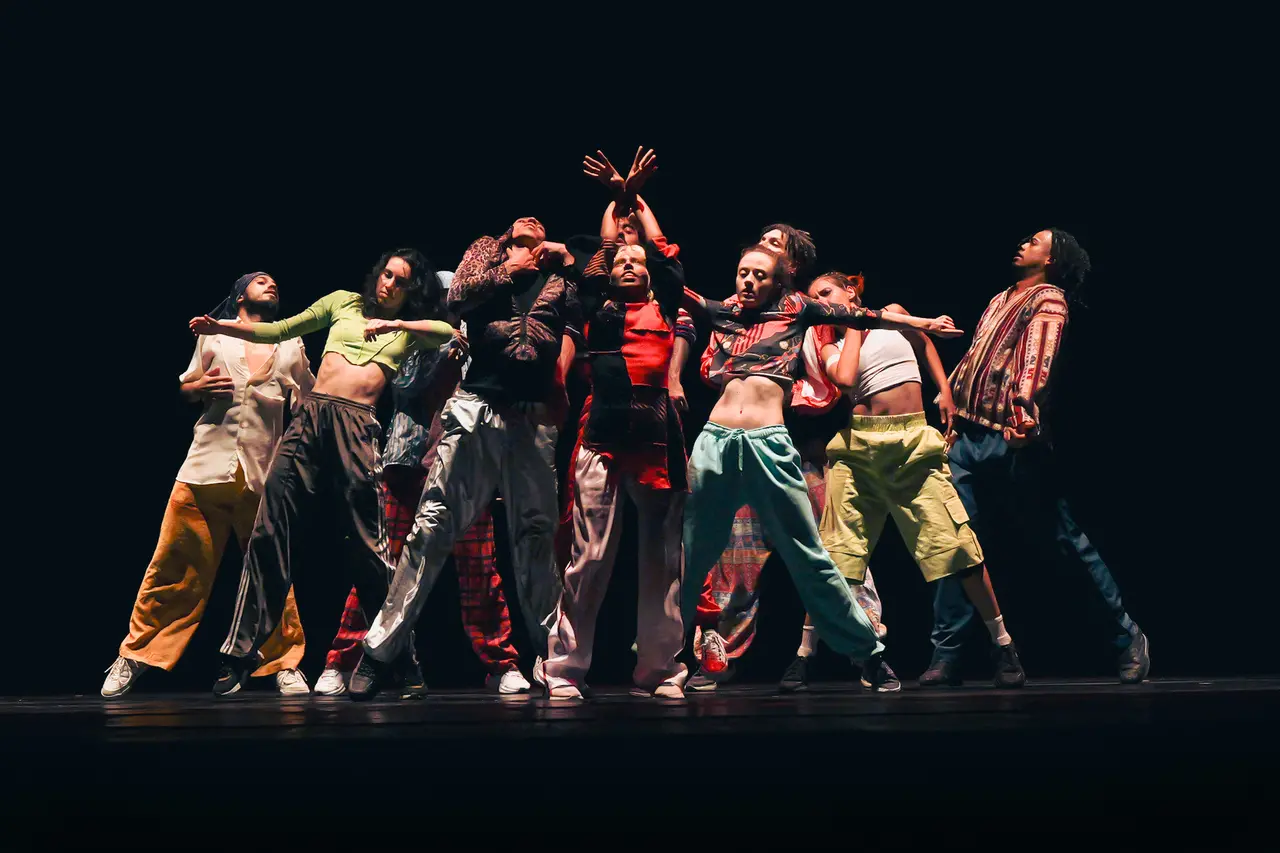 Imagem de divulgação do espetáculo 'Nosso Baile', com bailarinos reunidos ao centro num palco escuro.