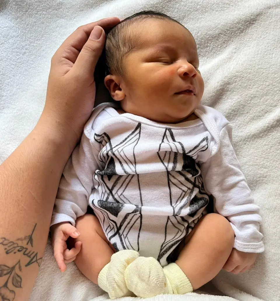 Bebê recém-nascido dormindo. Ele veste um body branco com traços do grafismo indígena.