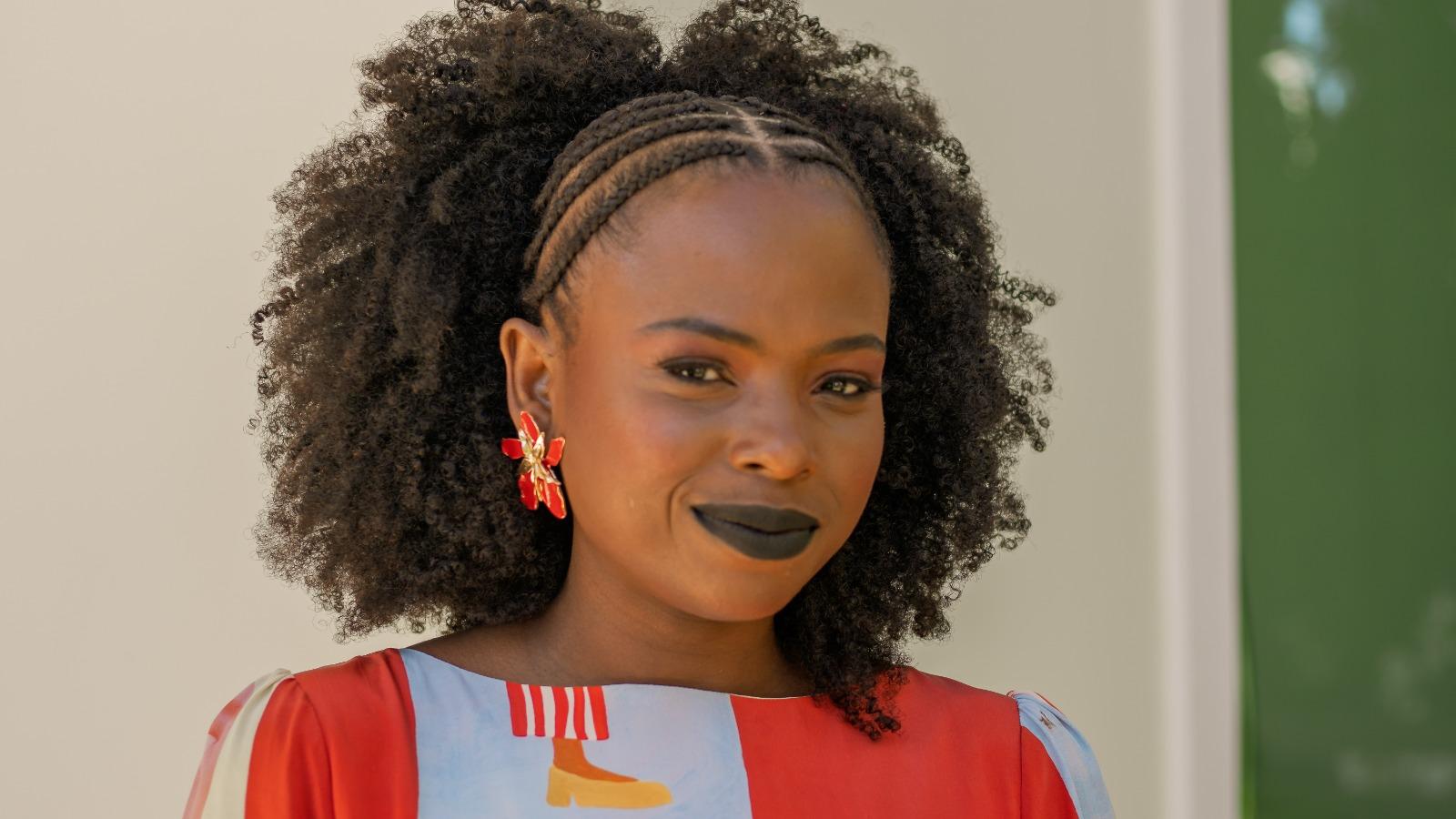 Mulher negra com cabelo afro e trançado na parte frontal, usando batom preto, vestido colorido e brincos vermelhos em formato de flor, posando em ambiente externo com fundo neutro.
