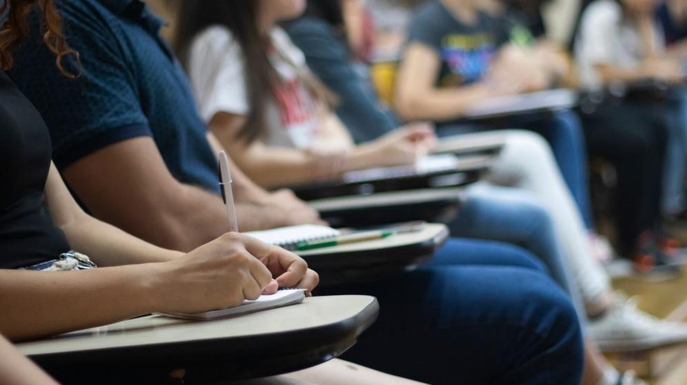 Na imagem, pessoas estão sentadas em uma sala de aula anotando em cadernos