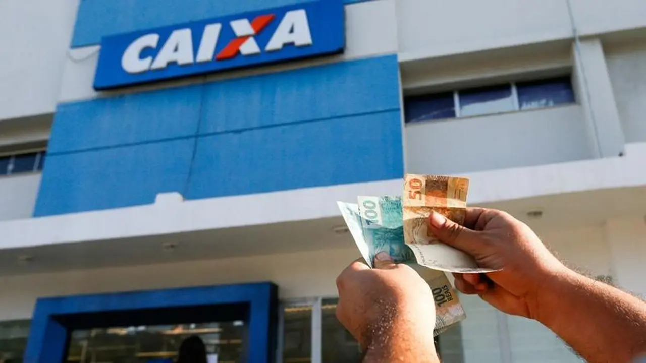 Pessoa segurando dinheiro em frente a uma agência da Caixa Econômica Federal, banco brasileiro com destaque na fachada.