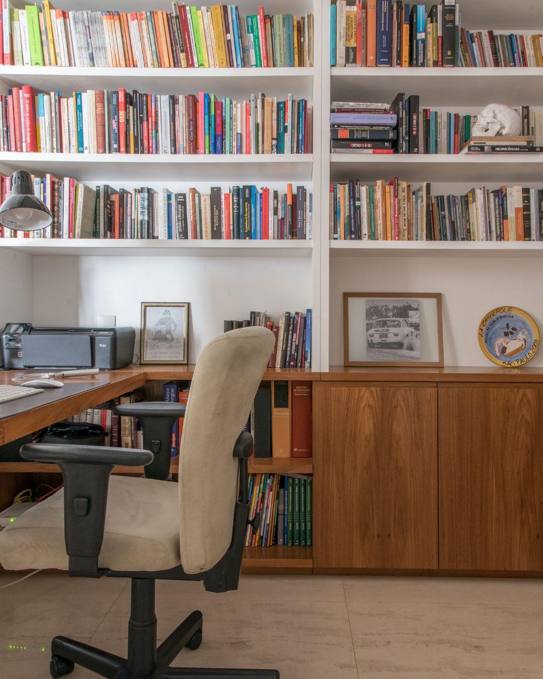 Estante de livros em um escritório com cadeira de escritório, mesa com impressora, fotos emolduradas e objetos decorativos, ambiente organizado para leitura e estudo.