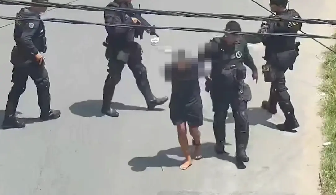 Policiais fortemente armados, vestidos com uniformes táticos, escoltam uma pessoa de roupas escuras e pés descalços por uma rua asfaltada sob o sol. O grupo caminha próximo ao meio-fio, e fios elétricos passam acima deles.