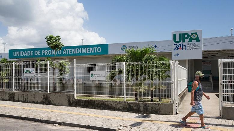 Esta imagem mostra a fachada de uma Unidade de Pronto Atendimento (UPA 24h), identificada por uma placa com o nome “Unidade de Pronto Atendimento” em destaque na cor verde. A estrutura é cercada por grades brancas, e há uma calçada pavimentada à frente. No canto direito da imagem, uma pessoa caminha pela calçada, usando boné, máscara, camiseta sem mangas e bermuda. O céu está parcialmente nublado e há árvores pequenas no entorno da unidade.