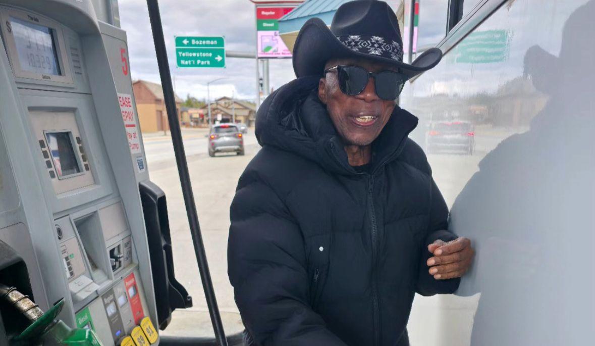 Milton Nascimento de chapéu preto e óculos escuros abastecendo o carro em um posto de gasolina dos Estados Unidos. Ele é um homem negro e idoso.