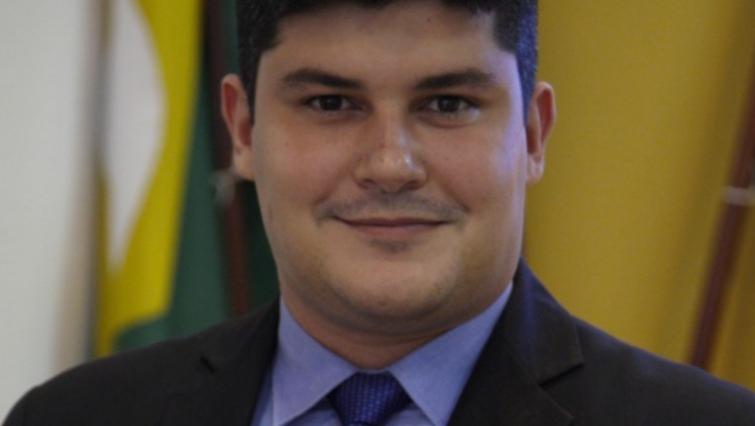 Joel Barroso é um jovem branco de cabelo preto bem cortado. Na foto, ele está com um leve bigode e usa terno preto com camisa social azul e gravata da mesma cor.