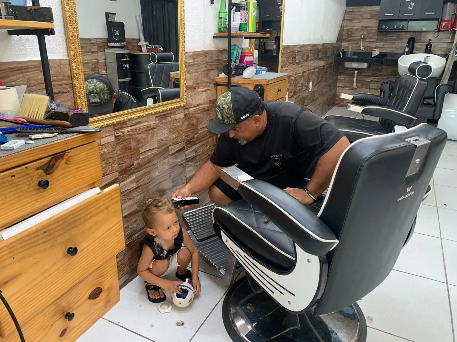 Na imagem, uma foto em um salão de cabeleireiro ou barbearia. Um homem barbudo com um boné escuro e uma camisa preta está agachado ao lado de uma cadeira de barbeiro grande e preta, segurando uma máquina de cortar cabelo. Ele está aparentemente interagindo com uma criança pequena, de cabelo claro e vestindo uma regata preta e branca e shorts brancos, que está agachada no chão na frente dele, segurando uma pequena bola. O fundo mostra uma parede com azulejos que imitam madeira, um espelho dourado refletindo a área, e móveis de madeira. O ambiente parece ser um salão de cabeleireiro ou barbearia em funcionamento, com equipamentos e produtos visíveis.
