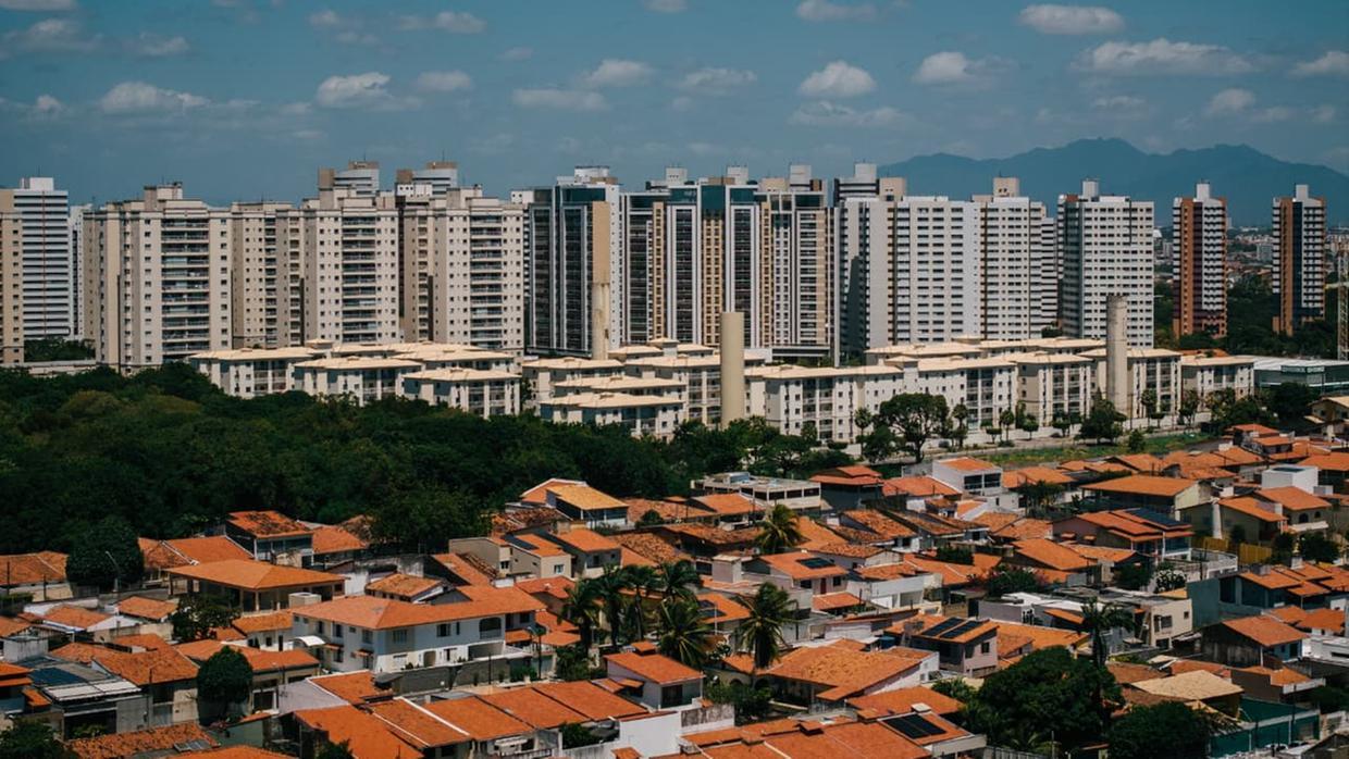 Foto panorâmica da cidade de Fortaleza, com diversos prédios altos em cores claras, predominantemente brancos, e, à frente, casas baixas com foco nos telhados avermelhados. Ao fundo, o céu azul limpo, com poucas nuvens brancas ao redor.