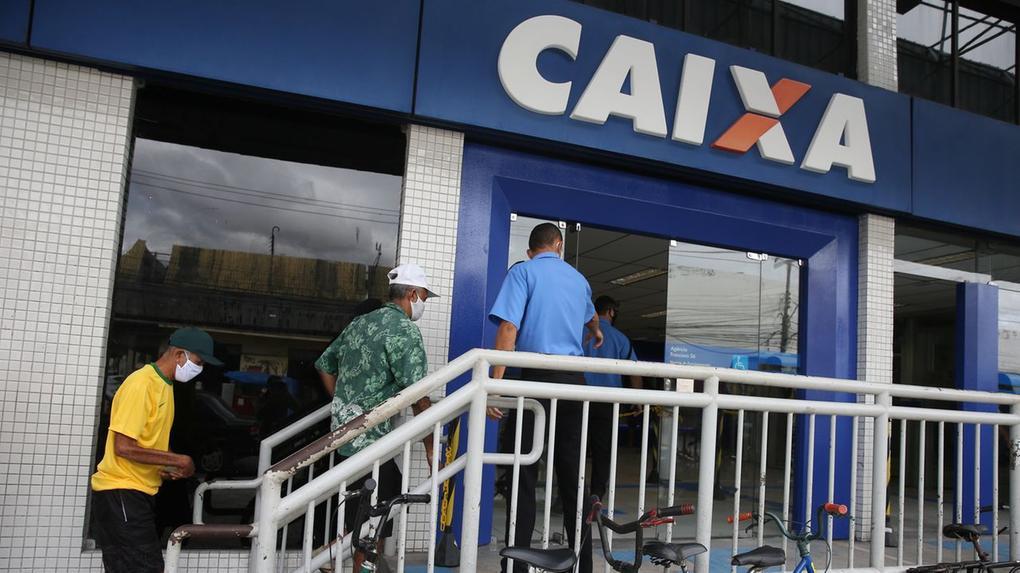 A foto mostra a fachada de uma sede da Caixa Econômica Federal, com a movimentação de clientes e um funcionário.