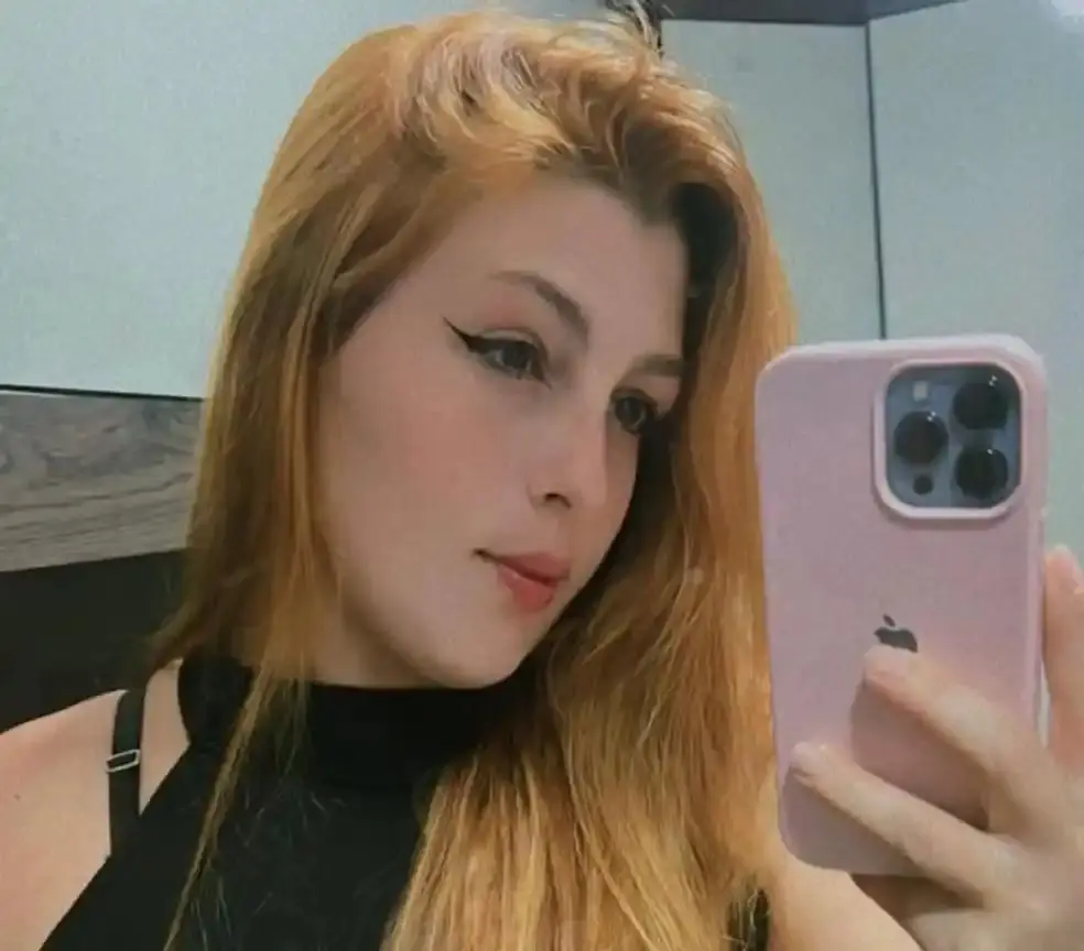 Jovem mulher com cabelo ruivo e maquiagem com delineado marcante, tirando uma selfie com smartphone rosa em ambiente moderno..