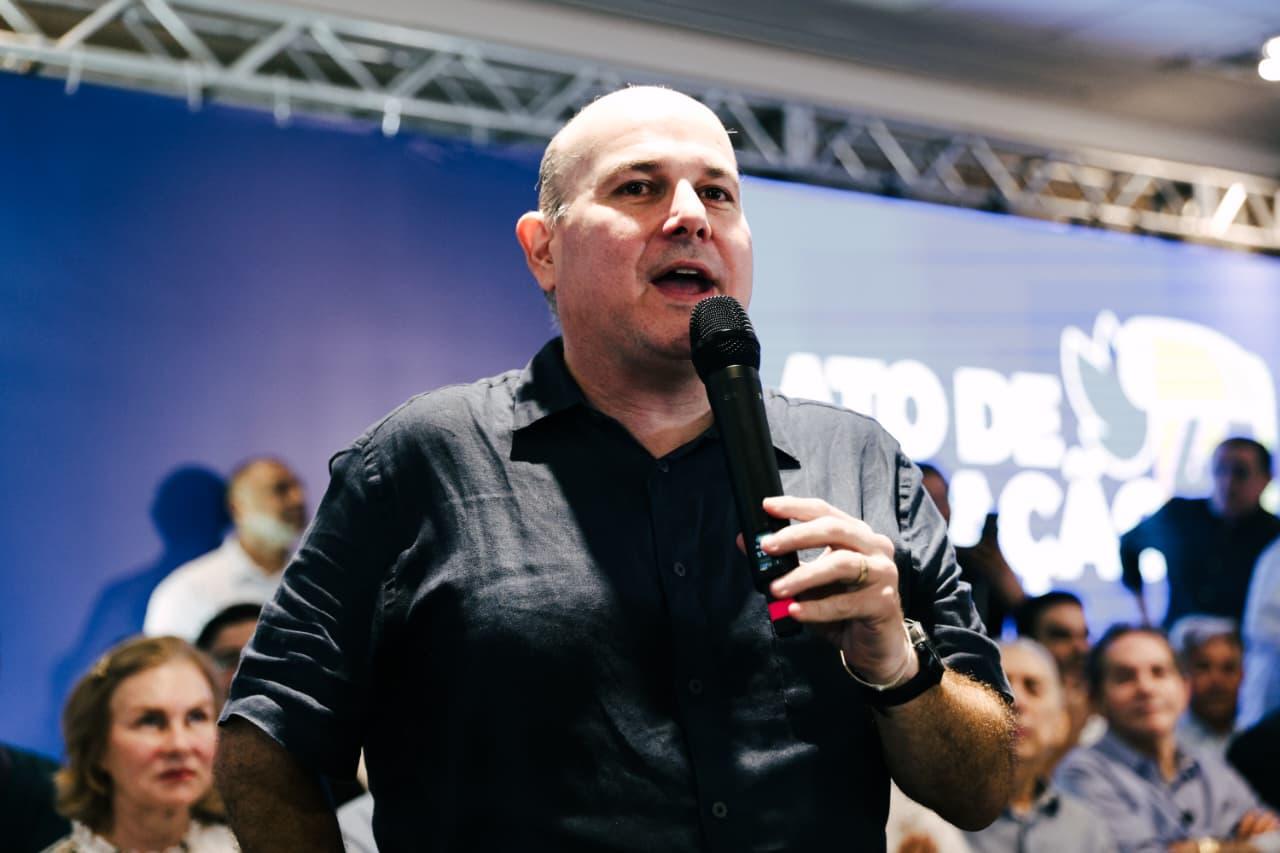 Roberto Cláudio, de camisa preta, com microfone na mão, discursando em palanque, com banner azul atrás.