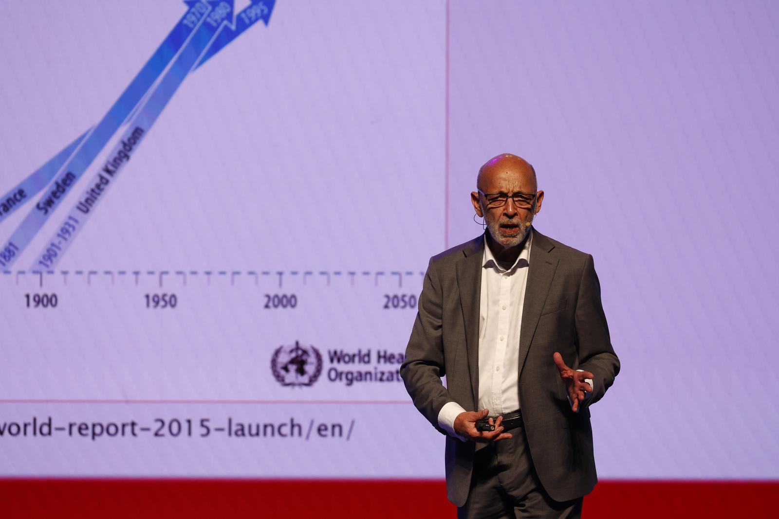 Imagem mostra o médico Alexandre Kalache durante palestra. Ele é um homem negro, careca, e veste camisa social branca por baixo de um terno completo cinza. Ele gesticula e atrás dele aparece um telão com gráficos.