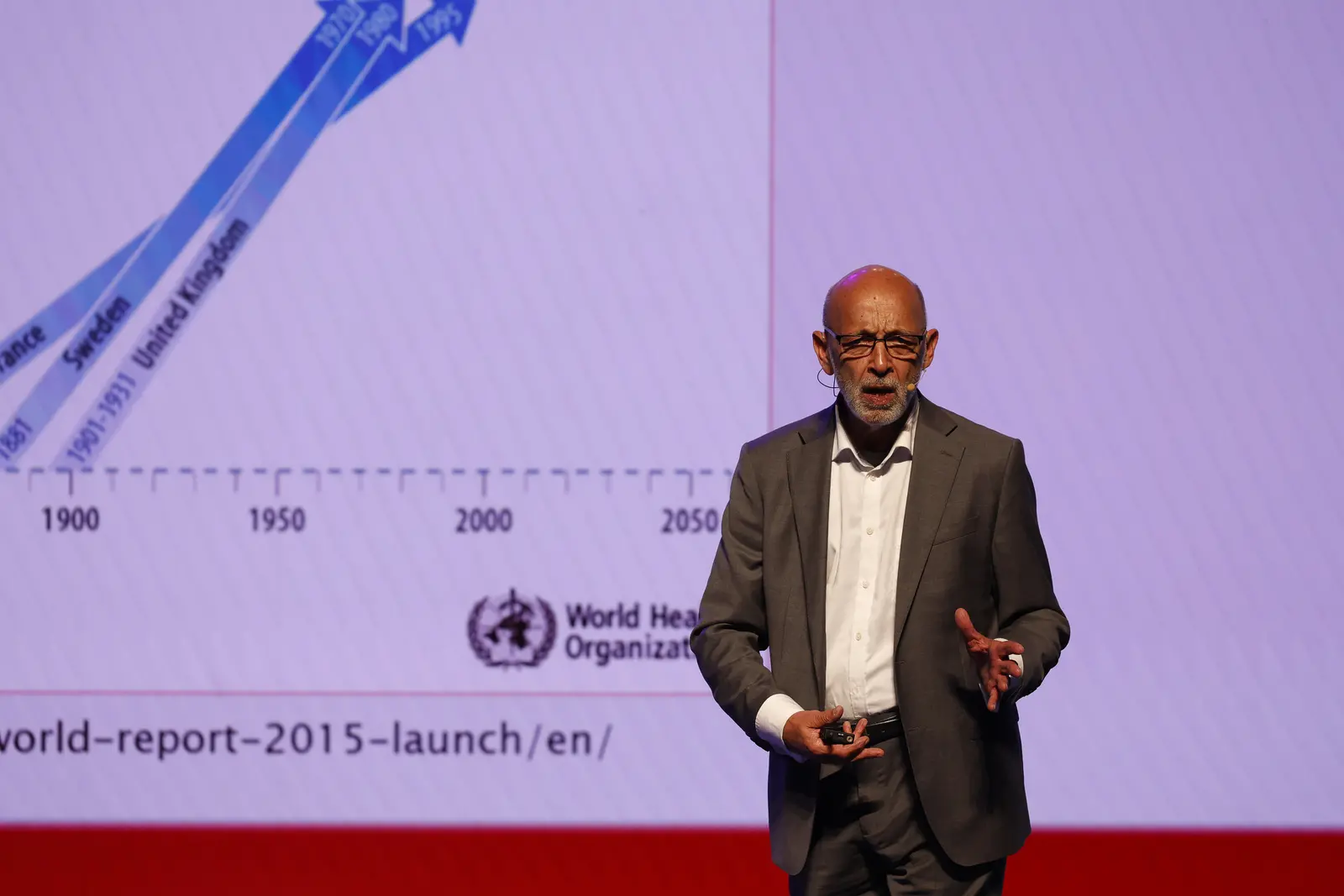 Imagem mostra o médico Alexandre Kalache durante palestra. Ele é um homem negro, careca, e veste camisa social branca por baixo de um terno completo cinza. Ele gesticula e atrás dele aparece um telão com gráficos.