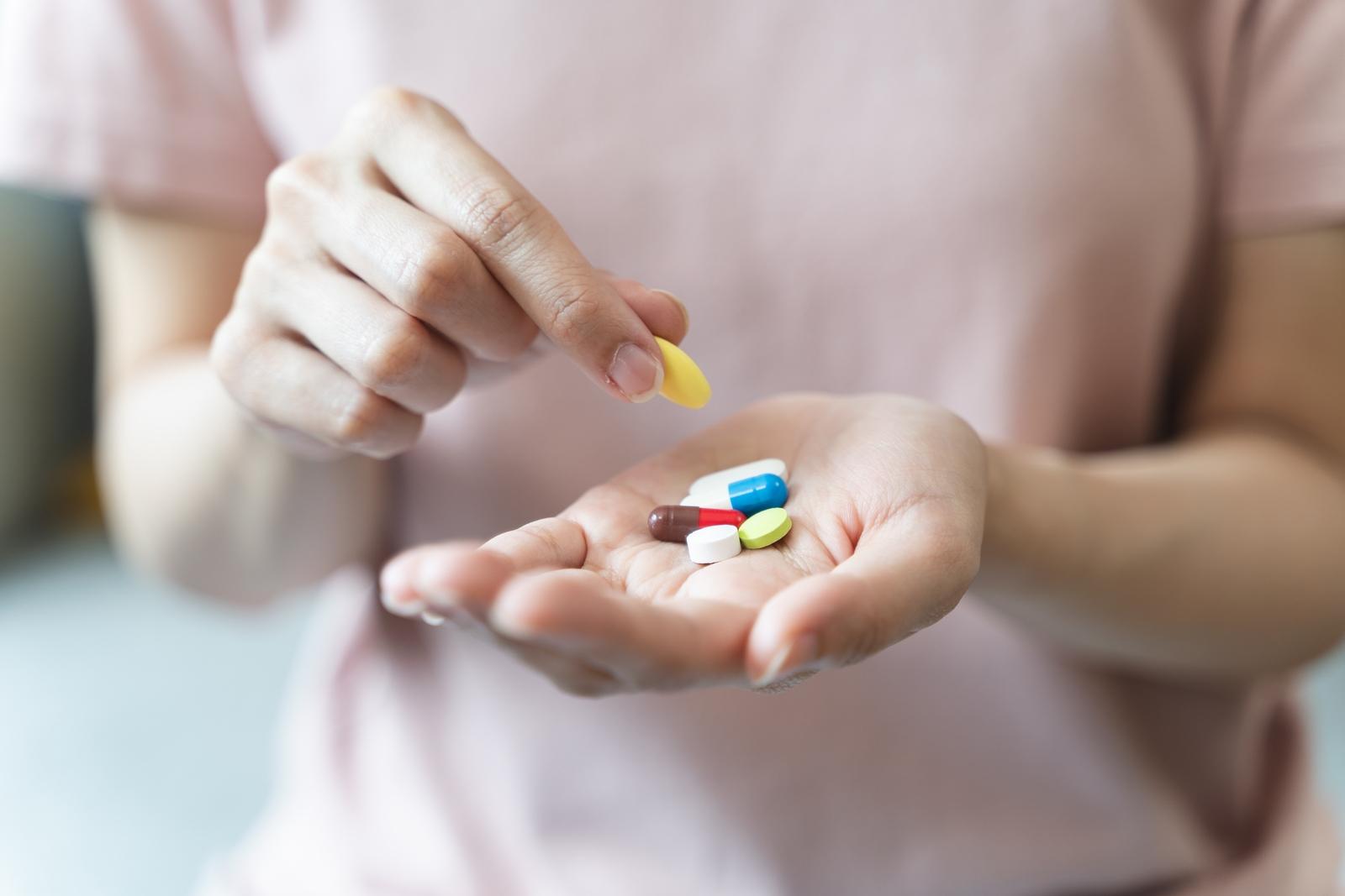 Imagem mostra mão de uma mulher segurando diversos comprimidos, enquanto a outra pega uma pílula amarela. A mulher veste uma camiseta rosa.