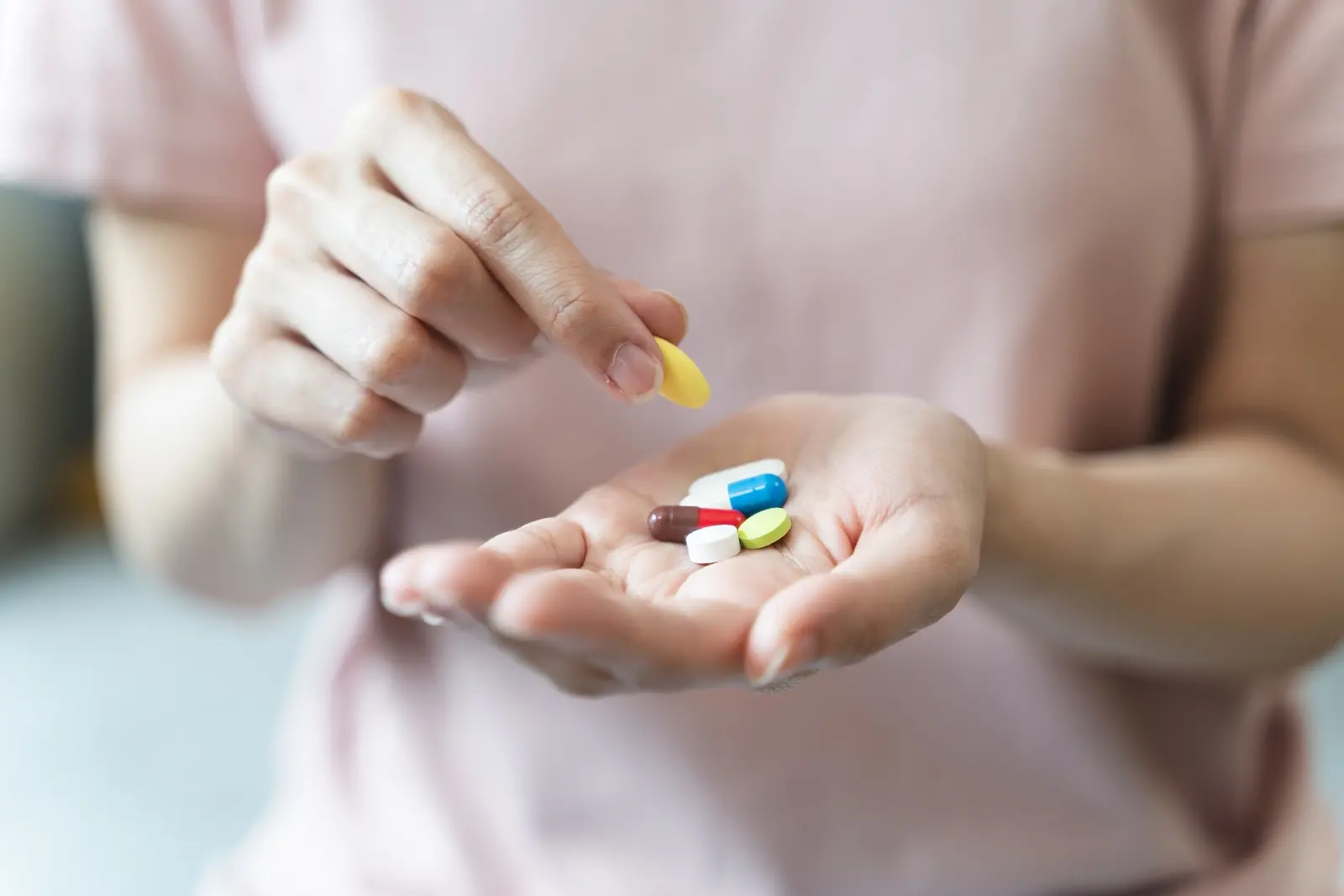 Imagem mostra mão de uma mulher segurando diversos comprimidos, enquanto a outra pega uma pílula amarela. A mulher veste uma camiseta rosa.