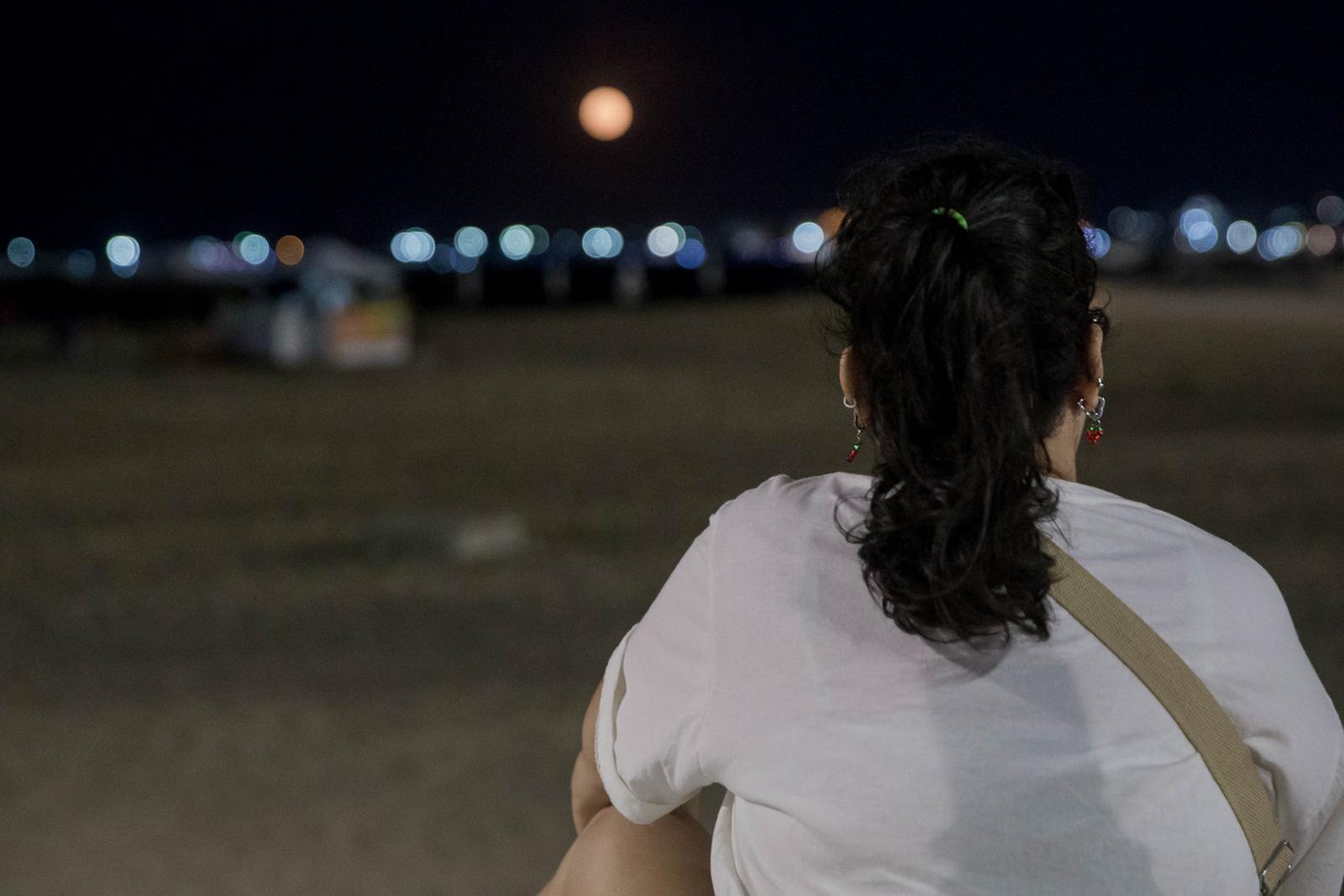 Imagem mostra mulher de costas, de cabelo preto e camiseta branca, olhando o mar durante a noite em Fortaleza.