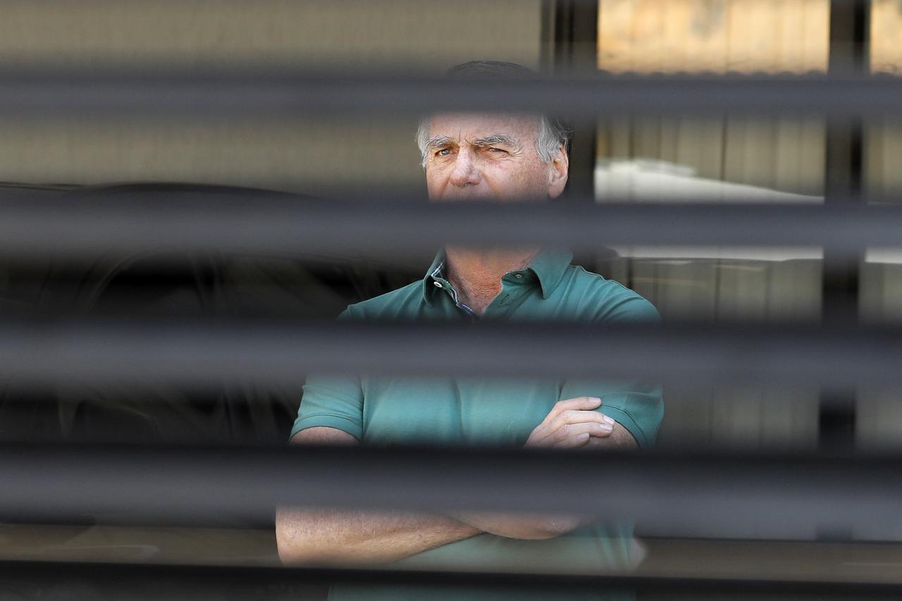 Imagem mostra o ex-presidente Jair Bolsonaro atrás de grades do portão de sua própria residência, em Brasília, onde cumpre prisão domiciliar. O ex-presidente é um homem idoso, de cabelos grisalhos, e está usando uma camisa de gola verde.