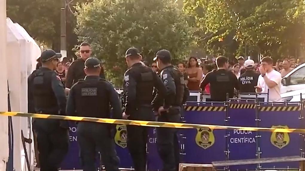 Polícia Civil atuando na cena de um incidente sob a sombra de árvores, com várias pessoas observando ao fundo, em frente ao cemitério.