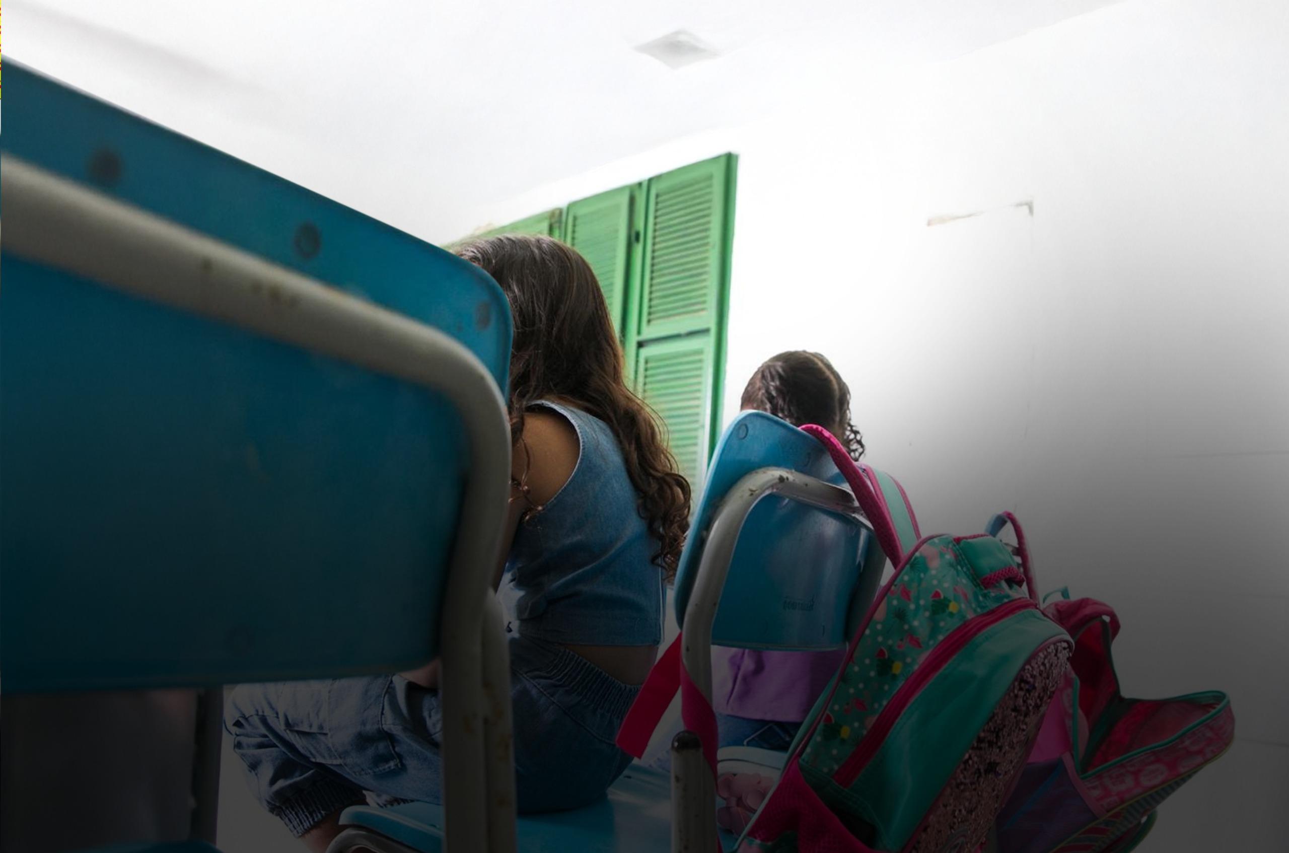 uas crianças sentadas em cadeiras escolares azuis, de costas, com mochilas coloridas penduradas nas cadeiras. A luz natural entra por uma janela próxima, iluminando o ambiente.