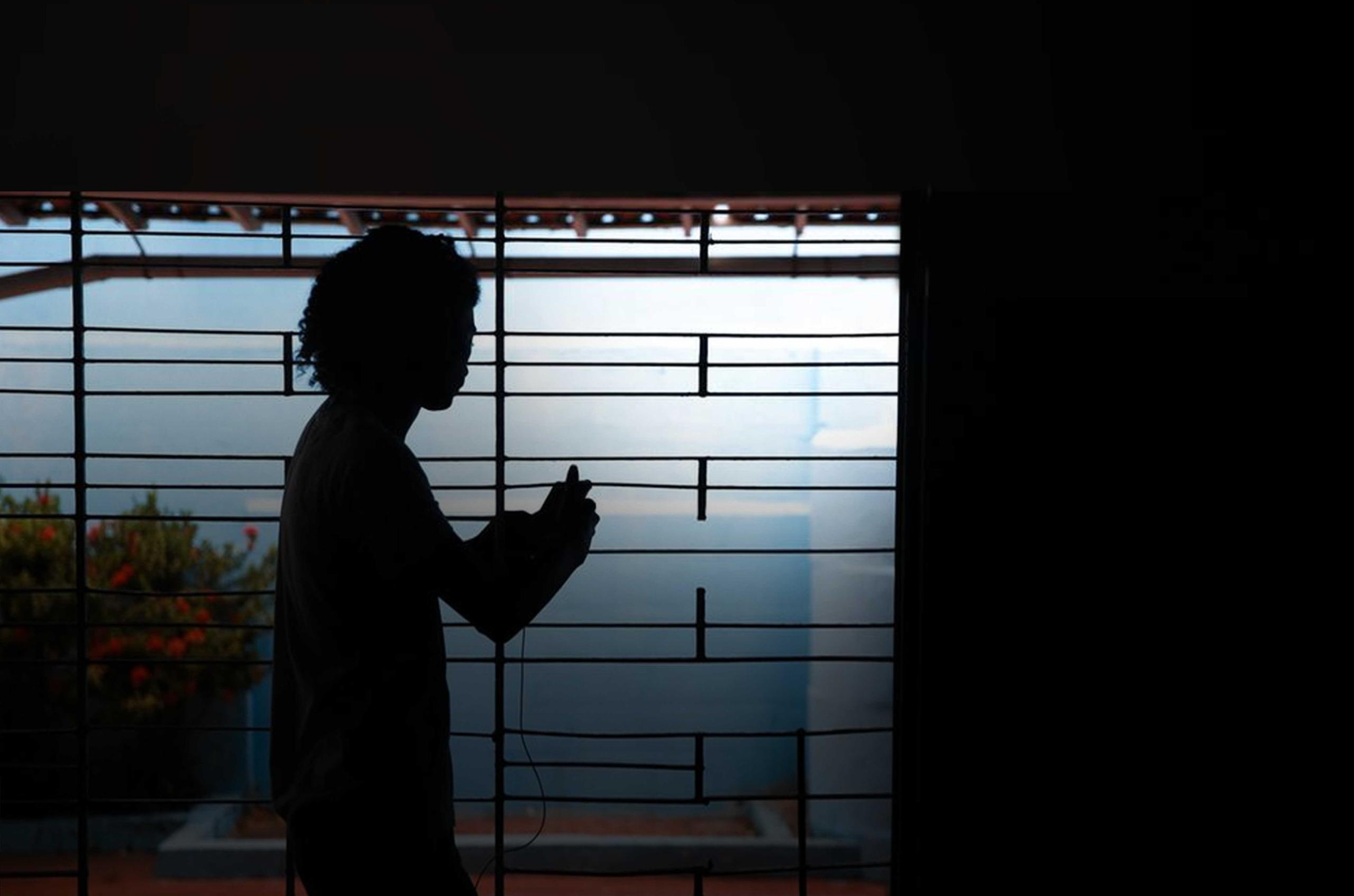 Silhueta de mulher em frente a uma grade, dentro de casa. Ao fundo, há um muro azul e uma planta com flores vermelhas. O clima é de reclusão e sofrimento.