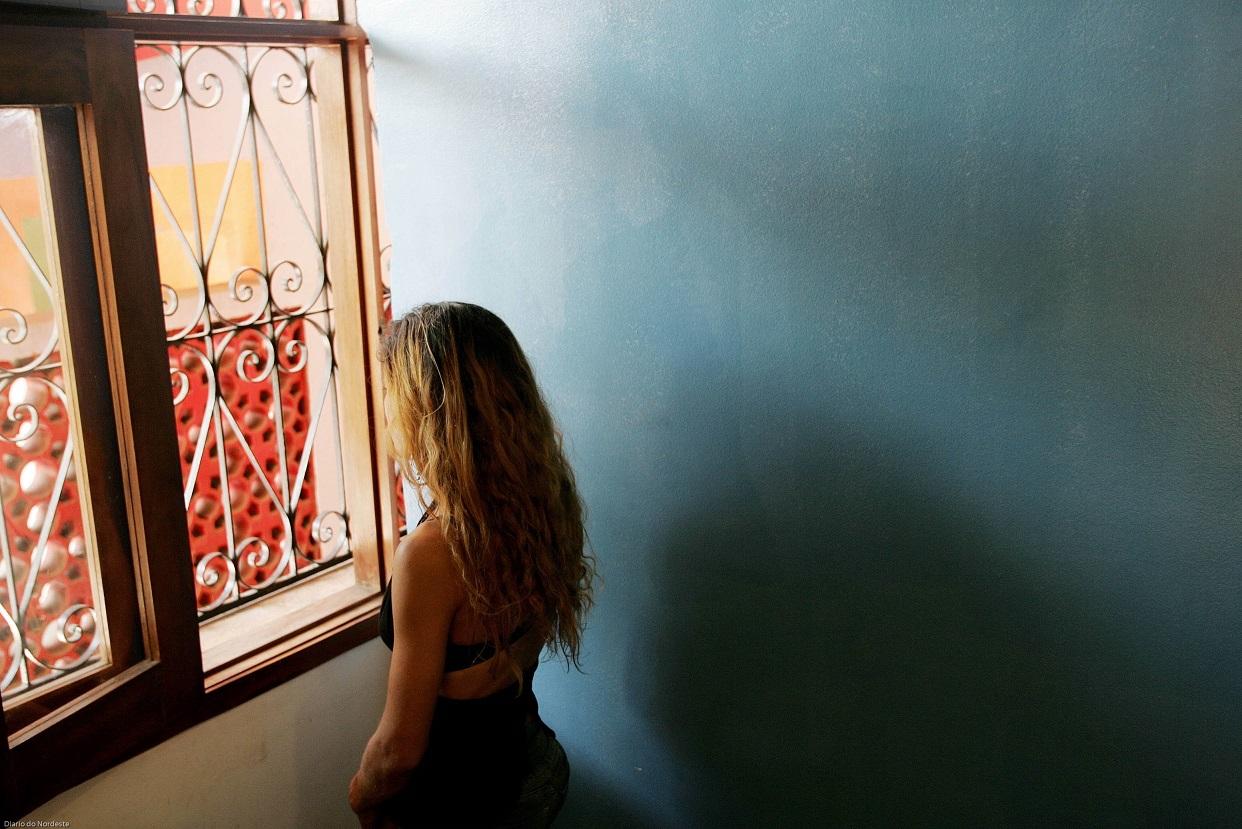 A imagem mostra uma jovem de cabelos longos e loiros, vista de costas, olhando pela janela de uma casa. Ela está sozinha em um ambiente interno com paredes em tons de azul.