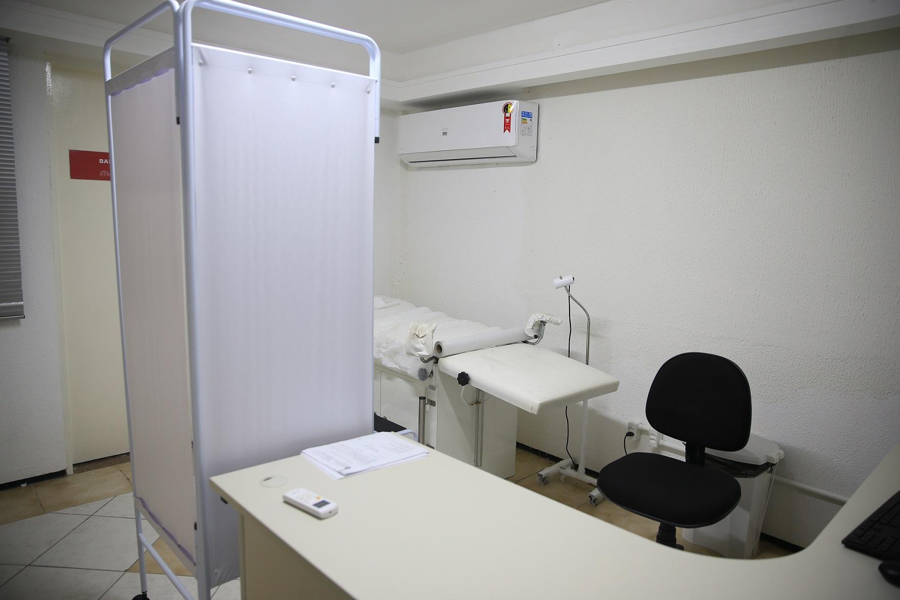 Sala de atendimento clínico, com divisória branca, maca, mesa de atendimento e cadeira. O ambiente é limpo, organizado e bem iluminado, sugerindo acolhimento e privacidade.