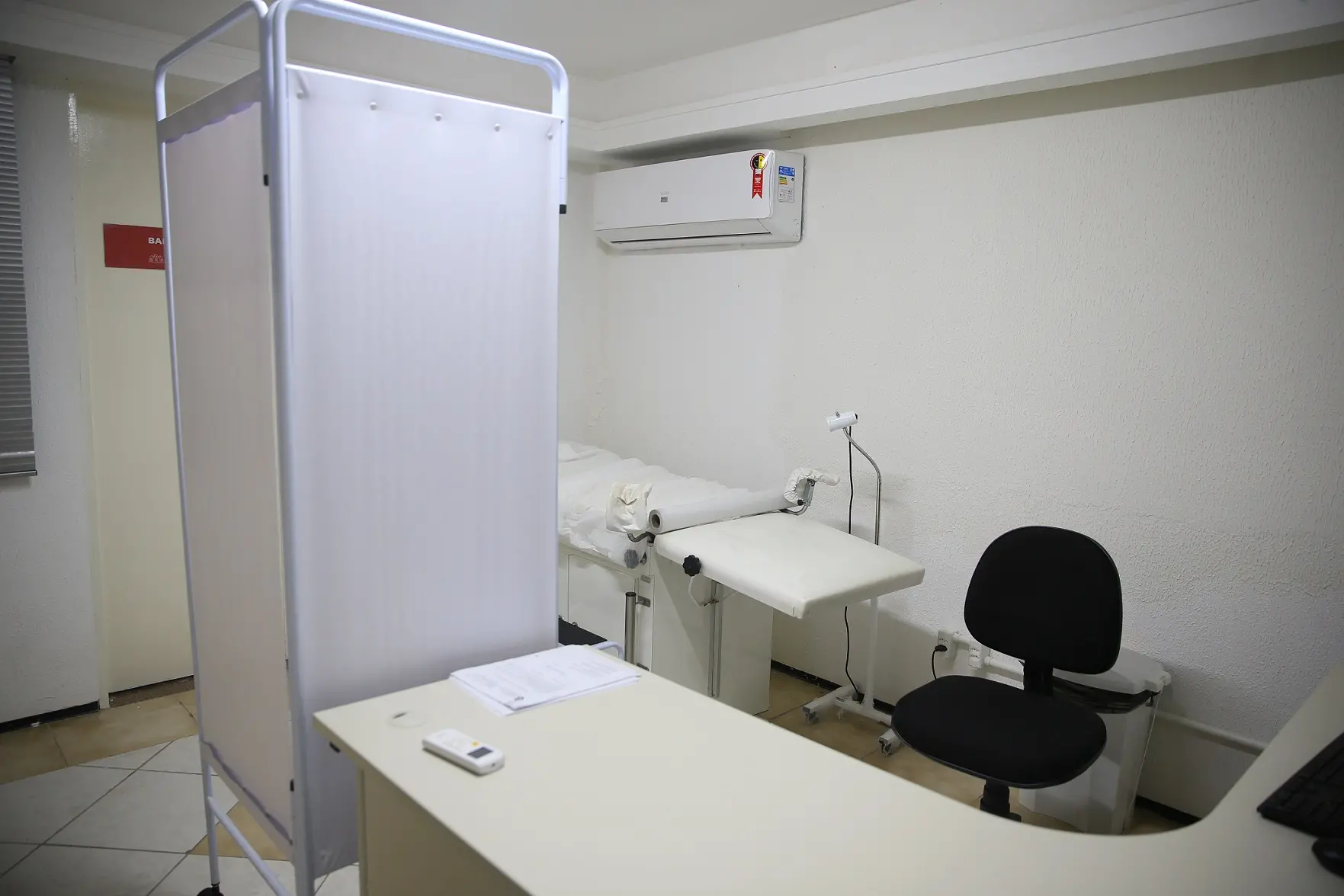 Sala de atendimento clínico, com divisória branca, maca, mesa de atendimento e cadeira. O ambiente é limpo, organizado e bem iluminado, sugerindo acolhimento e privacidade.