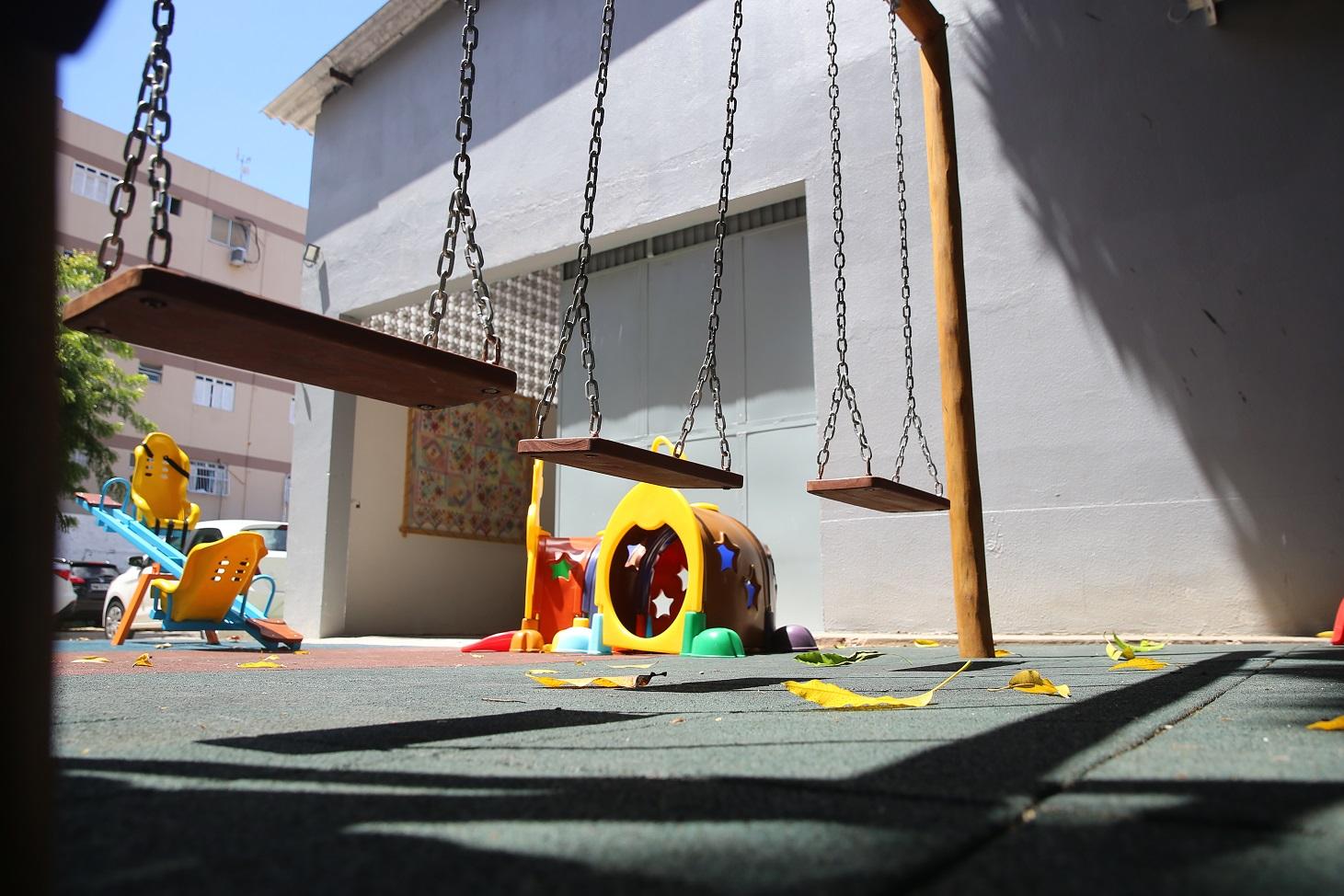 Pequeno parquinho infantil, com balanços de madeira pendurados por correntes e brinquedos coloridos ao fundo, como escorregadores e túneis. O chão é de piso emborrachado, e há folhas caídas espalhadas.
