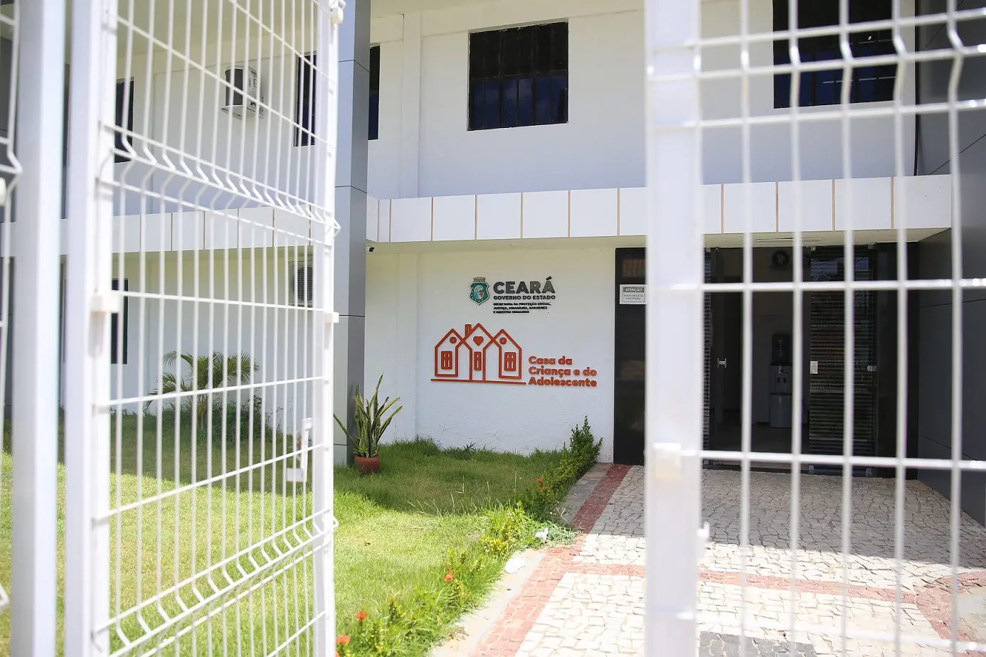 Fachada da Casa da Criança e do Adolescente, equipamento do Governo do Estado do Ceará em Fortaleza. O prédio é branco, com jardim verde e o logotipo institucional do governo e da unidade na parede.