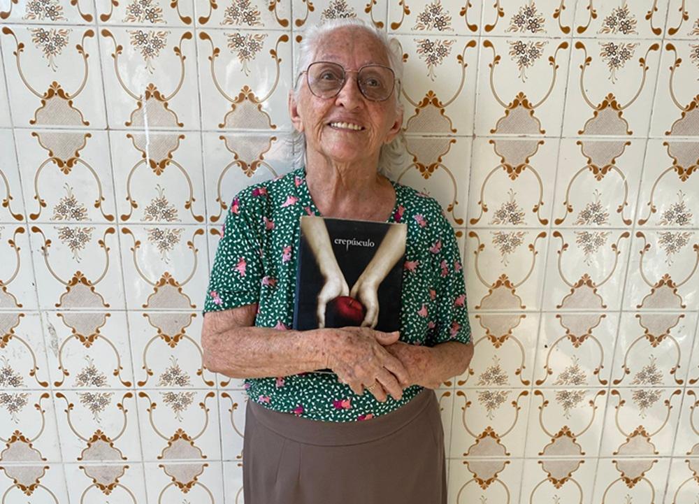 Na imagem, uma idosa, identificada na legenda como Dona Lourdinha Reis, de pele clara, cabelos brancos presos em um coque e óculos redondos, sorri enquanto segura um livro contra o peito com as duas mãos. Ela está vestindo uma blusa de manga curta com estampa floral verde e rosa sobre um fundo verde escuro e uma saia marrom de comprimento médio. O livro que ela segura é o primeiro volume da série Crepúsculo, com a capa que apresenta duas mãos segurando uma maçã vermelha. Ela está em pé na frente de uma parede com azulejos brancos e beges com um padrão floral repetitivo. A luz está clara, provavelmente natural.