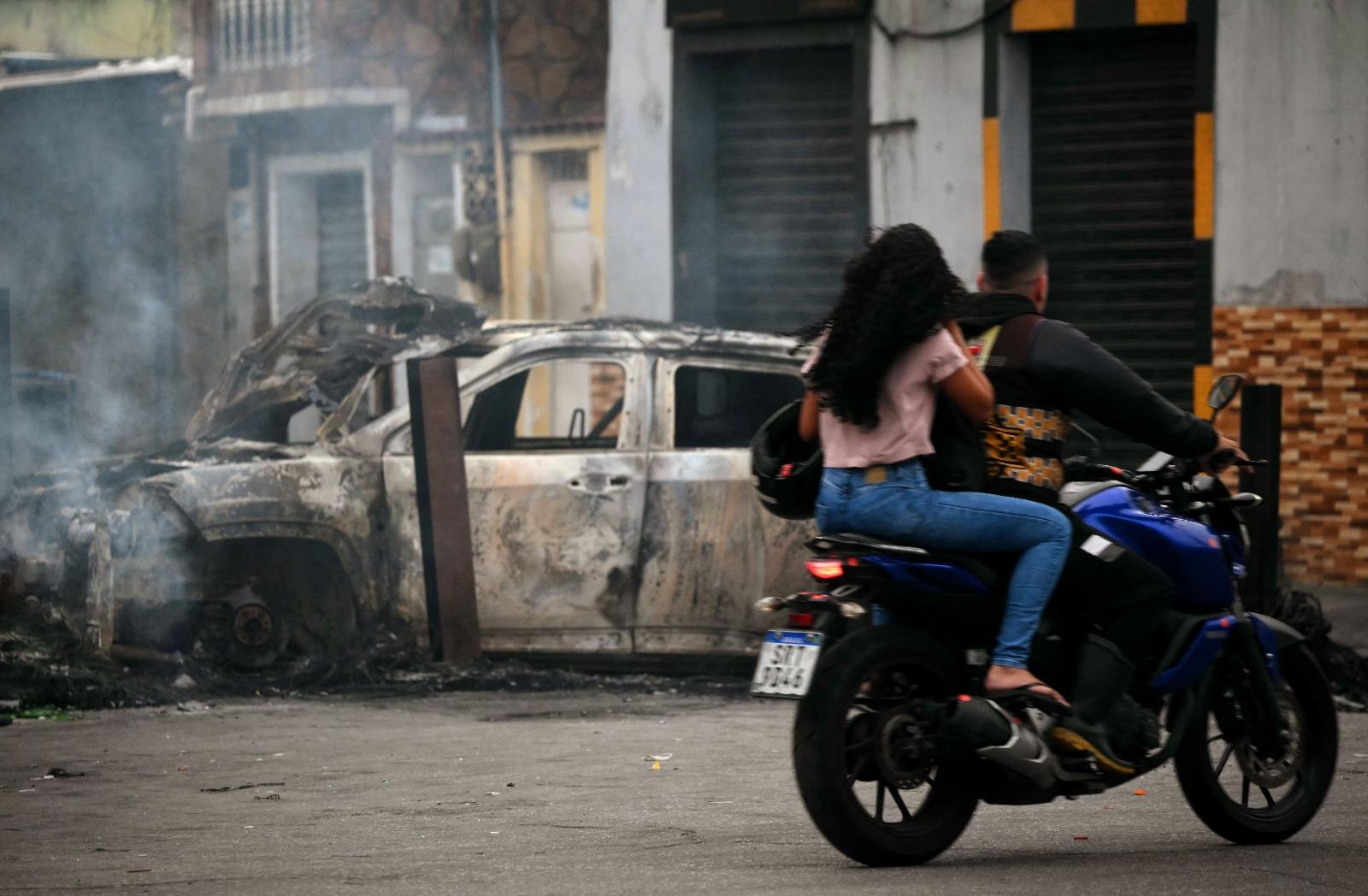 Casal de moto se depara com um carro incendiado servindo de barricada em uma rua.