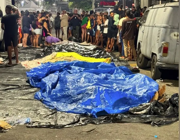 Imagem noturna mostrando dezenas de corpos cobertos por lonas plásticas (azul, amarela e pretas) enfileirados em uma rua escura. Em segundo plano, um grupo grande de moradores e curiosos está reunido, alguns olhando para os corpos, após uma operação policial na Penha, Rio de Janeiro. Uma Kombi branca está estacionada à direita.