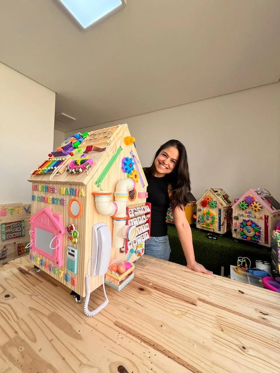 Inspirada pelo nascimento dos filhos, Paula Cruz passou a desenvolver diferentes tipos de painéis sensoriais. na foto, ela está atrás de uma casinha de madeira, que é um painel sensorial.