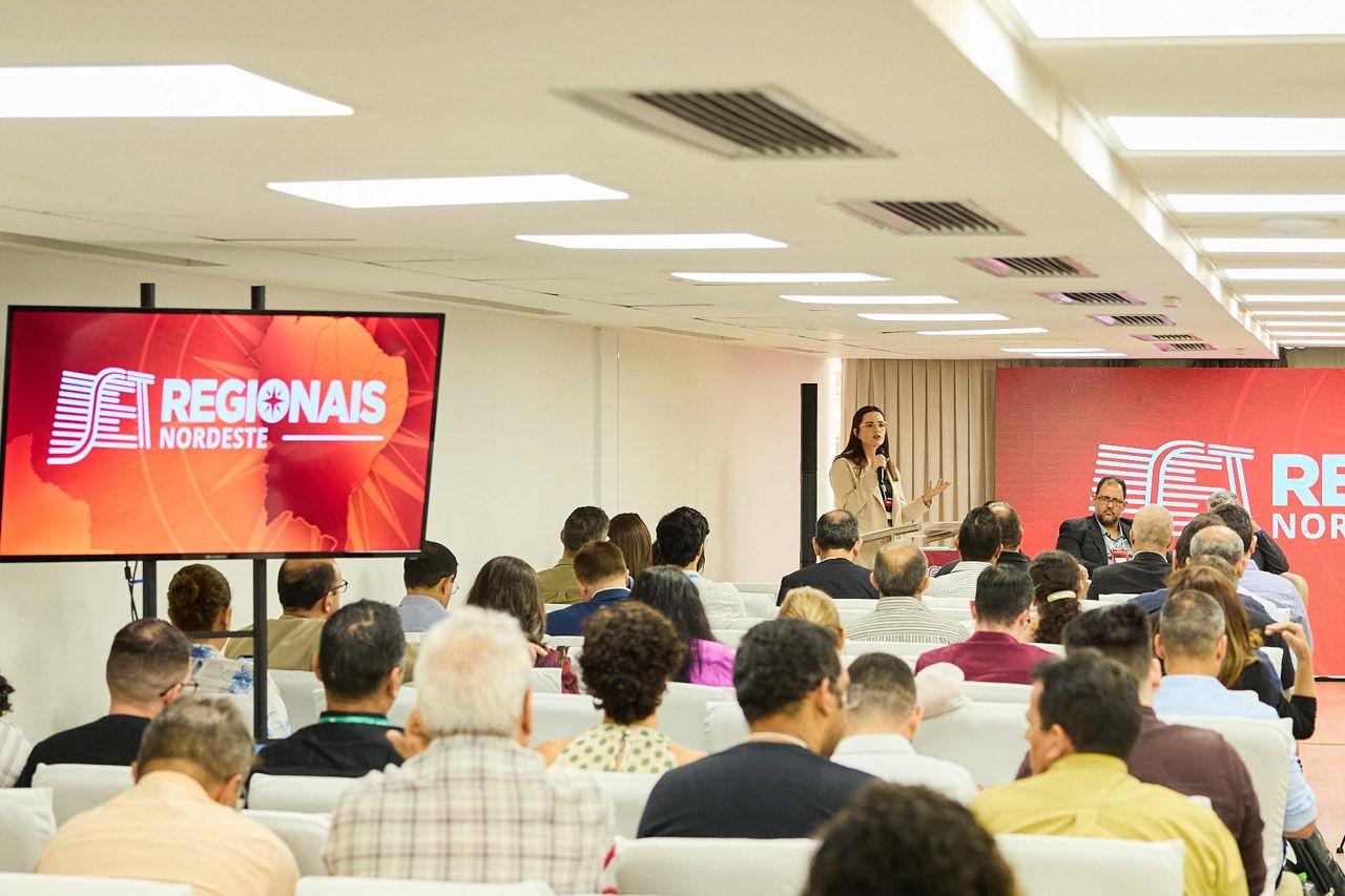 Imagem de uma conferência do Região Nordeste, com uma palestrante falando para uma audiência atentos, em ambiente corporativo bem iluminado.