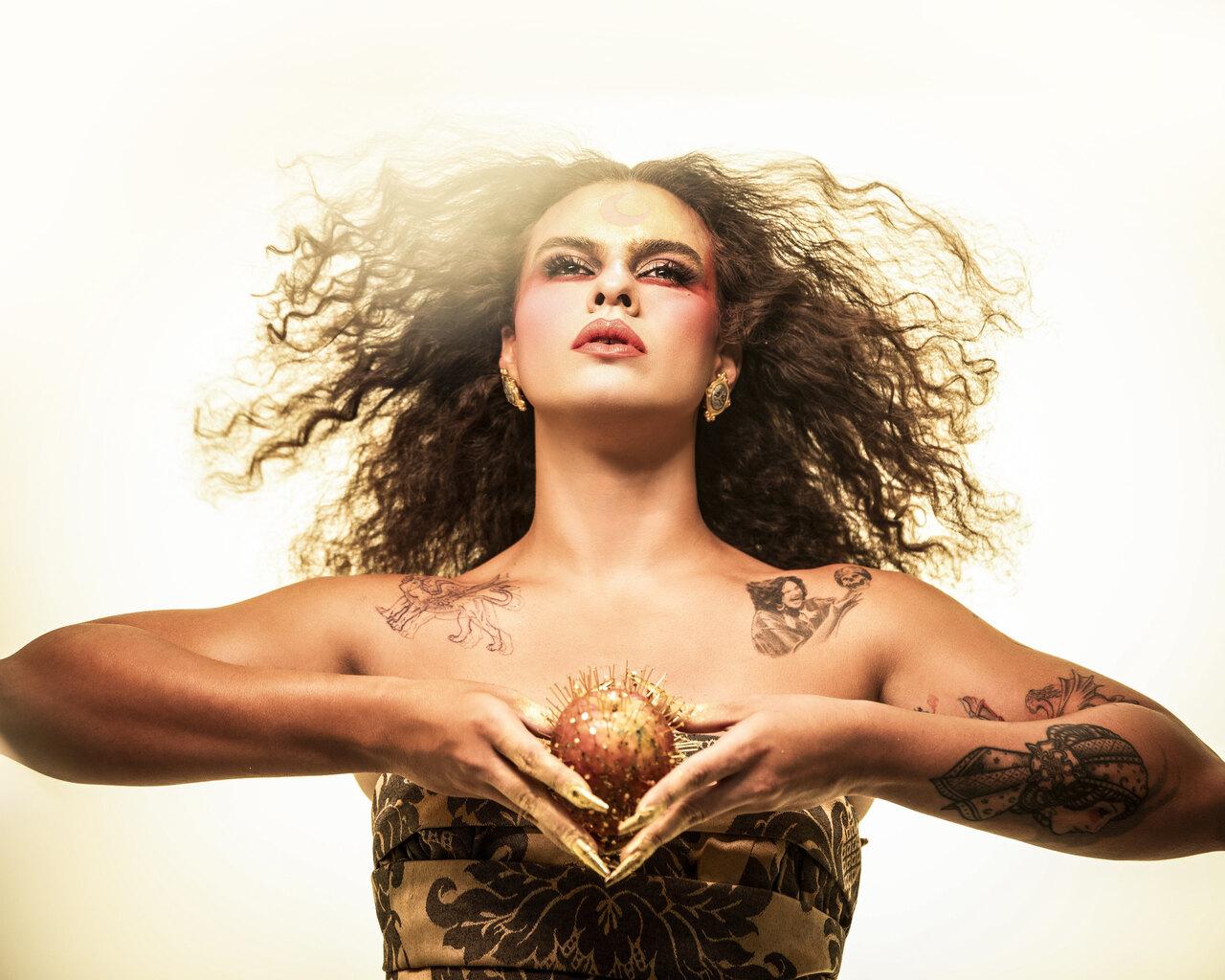 Foto da cantora Makem, uma mulher branca de cabelos castanhos cacheados e esvoaçantes. Ela segura uma maçã cheia de espinhos nas duas mãos.