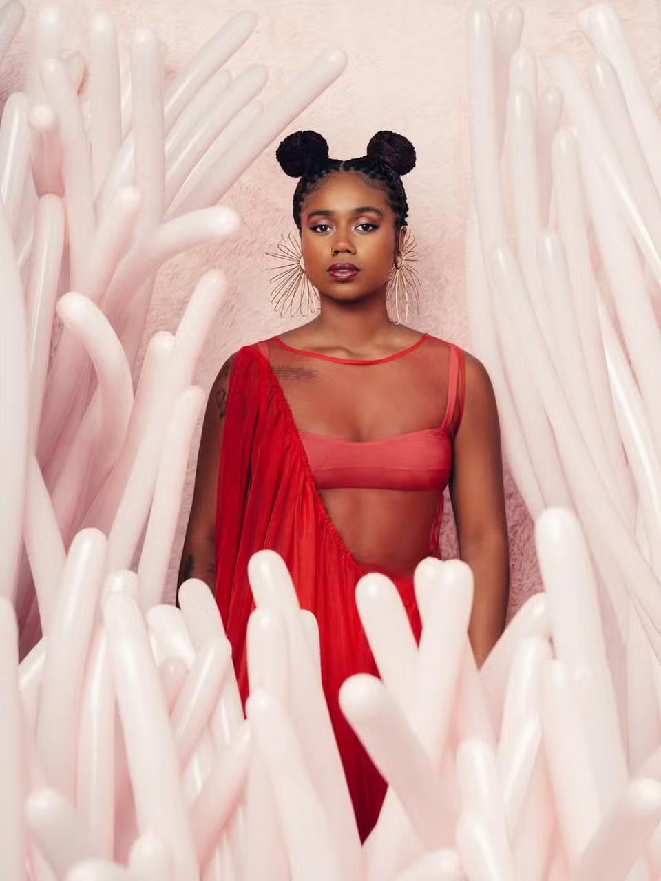 Melly é uma mulher negra e jovem com cabelos trançados e presos em dois coques no alto da cabeça. Ela usa um top rosa e um vestido com transparências vermelhas e posa séria em um cenário cheio de balões de cor clara.