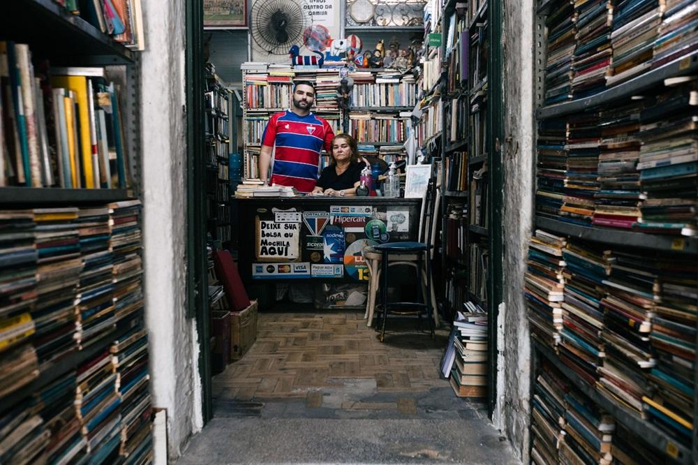 Na imagem, uma tomada em plano médio, através de uma porta ou corredor estreito, que mostra o interior de um sebo (livraria de livros usados). As laterais da imagem são dominadas por estantes altas e abarrotadas de livros, criando um túnel visual. No centro, ao fundo, atrás de um balcão, estão duas pessoas: um homem e uma mulher. O homem, em pé e ligeiramente atrás, usa uma camisa polo azul e vermelha. A mulher, sentada ou inclinada, usa uma blusa preta. Ambos olham diretamente para a câmera. O balcão à frente deles tem adesivos de cartões de crédito e um cartaz que diz, em parte, 